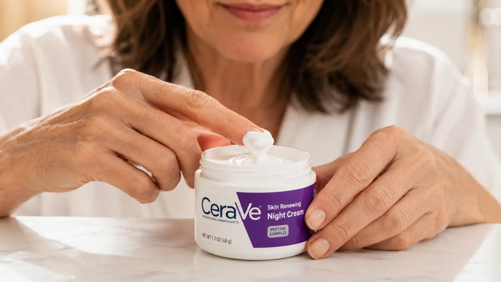 Hand scooping a small amount of CeraVe Skin Renewing Night Cream from the jar, white cream texture visible