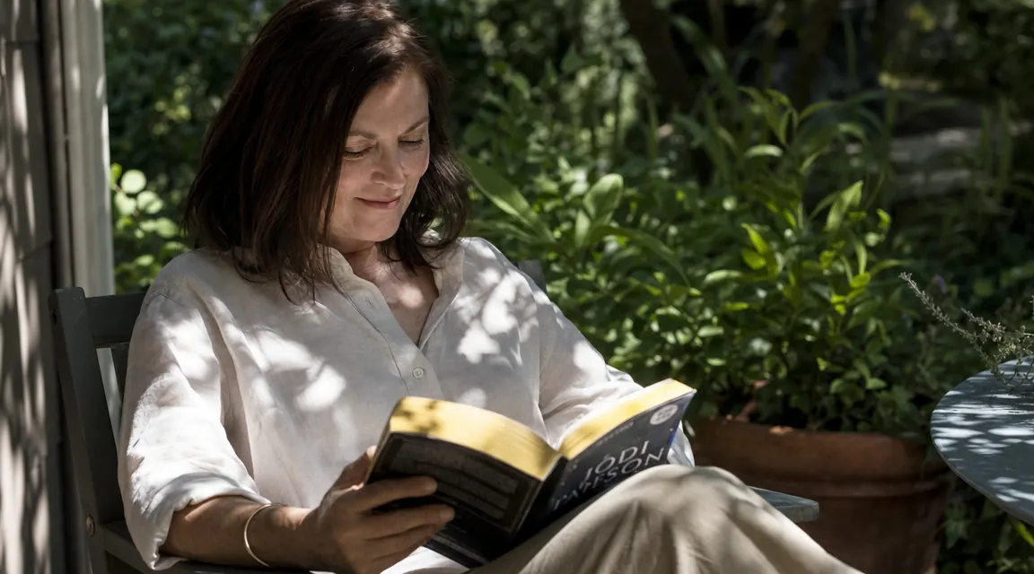 Outdoor scene of a woman in her 50s sitting in a garden or on a porch, reading a book in soft midday sun