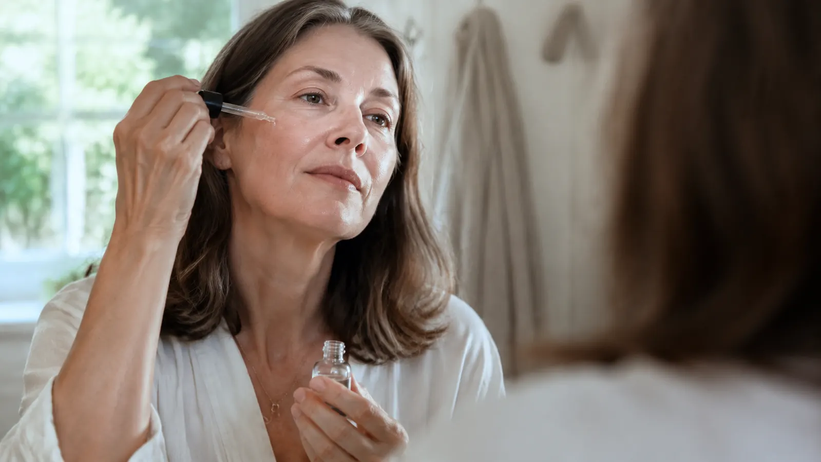Woman in her 50s applying serum to her face in front of a bathroom mirror, morning light, relaxed expression
